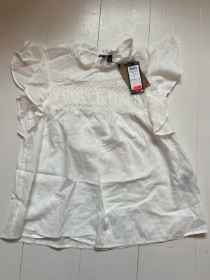 Vit blus med spets och volang - Söt vit blus från vero Moda med broderade prickar, spetsdetaljer över bröstet och volang på axlarna. Blusen har korta ärmar och rund halsringning. Materialet är lätt och luftigt, perfekt för en fräsch look.