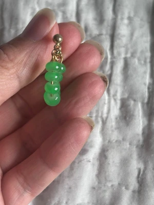 Gröna pärlörhängen i guldton - Säljer ett par örhängen med gröna, runda pärlor trädda på en båge med guldfärgade detaljer. Örhängena har en modern och lekfull design som sticker ut. Perfekta för dig som vill addera färg till din stil.