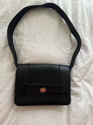 Svart axelväska med gulddetalj - Stilren svart axelväska i slätt konstläder med bred axelrem och rektangulär form. Väskan har ett lock med guldfärgat vridlås framtill som ger en snygg touch. Perfekt för dig som gillar minimalistisk design och vill ha plats för det viktigaste.