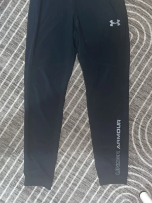 Svarta träningsbyxor Under Armour - Säljer ett par svarta träningsbyxor från Under Armour med logga på låret och text längs benet. Byxorna har smal passform och är tillverkade i ett stretchigt funktionsmaterial som passar perfekt till gym eller löpning.