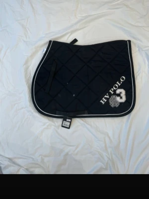 Svart schabrak HV Polo - Säljer ett svart schabrak från HV Polo med vit kant och broderad logga samt siffran 3. Schabraket har rutmönster och är tillverkat i slitstarkt tyg, perfekt för dig som gillar hästsport och vill ha en snygg detalj till din utrustning. Storlek cob/full