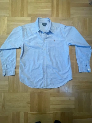Ljusblå skjorta från Ralph Lauren - Klassisk ljusblå skjorta från Ralph Lauren med broderad logga på bröstet. Skjortan har lång ärm, knappar framtill och en tidlös passform. Perfekt till jeans eller chinos för en clean och stilren look. Storlek S-M men kan skicka mått.