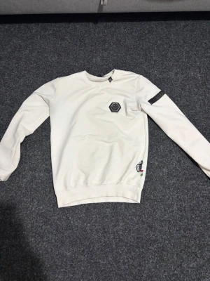 Vit sweatshirt från Philipp Plein - Snygg vit sweatshirt från Philipp Plein med rund halsringning och ribbade muddar. Tröjan har ett svart PP-logomärke på bröstet och en detalj på ärmen. Tillverkad i mjukt bomullsmaterial för en bekväm känsla.