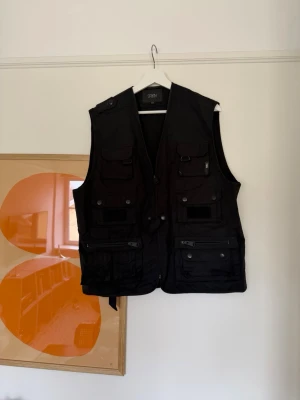 Svart utility-väst från Drkn - Utility-väst i svart från Drkn med flera fickor framtill, dragkedjor och tryckknappar. Västen har en rak passform och är tillverkad i ett slitstarkt tyg, perfekt för dig som gillar streetwear och funktionella detaljer.