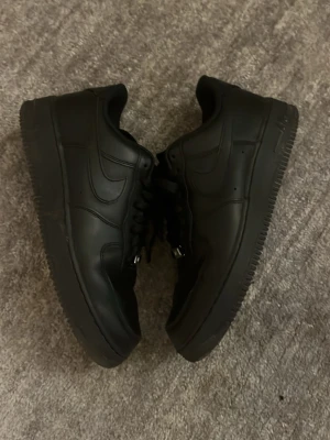 Svarta Nike Air Force 1 sneakers - Säljer ett par helsvarta Nike Air Force 1 sneakers i läder. Klassisk låg modell med svart sula, svarta skosnören och den ikoniska swoosh-loggan på sidan. Perfekta för dig som gillar stilrena och tidlösa sneakers.