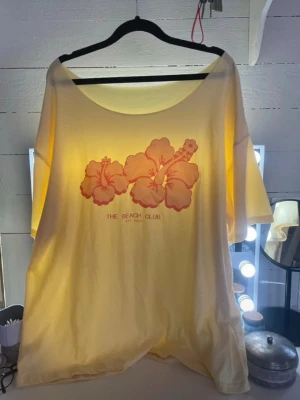 Gul t-shirt  - Snygg gul t-shirt från The Beach Club med stort blommotiv i orange och text framtill. T-shirten har korta ärmar, rund halsringning och en loose passform. Perfekt för sommarens alla dagar och enkel att styla till jeans eller shorts.