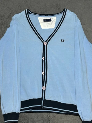 Fred Perry Cardigan Light Blue – Laurel Wreath Logo – XXL - Stilren Fred Perry cardigan i ljusblått med klassiska kontrastdetaljer i navy och vitt samt broderad laurel wreath-logo på bröstet.  Tunn stickning i bomull som passar perfekt för lager på lager och en klassisk brittisk stil.  Detaljer • Fred Perry • Ljusblå cardigan • Laurel wreath logo • Kontrastdetaljer vid krage och nederkant • Knäppning framtill • Storlek XXL • 100% bomull  Bra begagnat skick.  📦 Skickas snabbt 💬 Hör av dig vid frågor