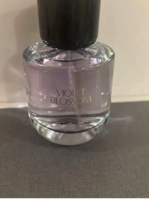 Zara Violet Blossom parfym - En elegant parfymflaska från Zara med doften Violet Blossom. Flaskan är rundad i genomskinligt glas med en svag lila ton och har ett svart lock. Texten på flaskan är i guld. Perfekt för dig som gillar blommiga och fräscha dofter.