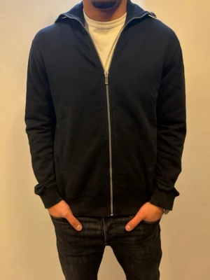 Svart hoodie med dragkedja från J.Lindeberg - Svart hoodie från J.Lindeberg med hel dragkedja framtill och ribbade muddar. Stort broderat J.Lindeberg-logotyp på ryggen. Tillverkad i mjukt bomullsmaterial och har en klassisk huva. Perfekt för en avslappnad och stilren look.