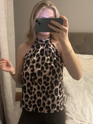Leopardmönstrad halterneck topp - Snygg halterneck topp med leopardmönster i svart och beige. Toppen är ärmlös och har en hög halsringning som knyts i nacken. 