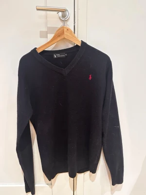 Svart stickad tröja från Polo Ralph Lauren - Klassisk svart stickad tröja från Polo Ralph Lauren med V-ringning och den ikoniska röda logotypen broderad på bröstet. Tröjan har långa ärmar och ribbade muddar vid ärmslut och nederkant. Perfekt för lager på lager och enkel att matcha.