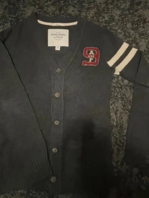 Mörkgrå cardigan från Abercrombie & Fitch - Collegeinspirerad mörkgrå cardigan med knappar framtill och vita ränder på ena ärmen. Broderad röd patch med texten 'A and F' på bröstet. Klassisk v-ringad modell som passar perfekt till en avslappnad stil.