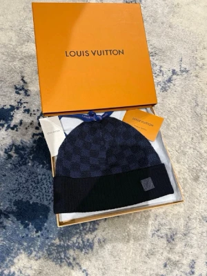 Mörkblå Louis Vuitton mössa - Snygg mörkblå mössa från Louis Vuitton med klassiskt rutmönster och vikt kant. Mössan har en diskret LV-logga på kanten och är tillverkad i mjukt, stickat material som känns lyxigt och bekvämt. Perfekt accessoar för att lyfta din stil.