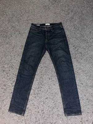 Jack & Jones  - Snygga mörkblå jeans från Jack & Jones med klassisk rak passform och fem fickor. Jeansen har kontrastsömmar och en läderpatch bak i midjan. Tillverkade i slitstark denim, perfekta för en clean och tidlös look.