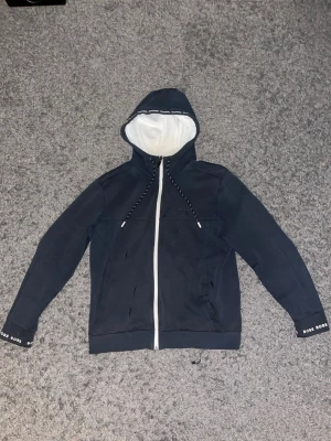 Hugo boss hoodie - Snygg mörkblå hoodie från BOSS med vit dragkedja och vit insida i huvan. Hoodien har dragsko med svarta och vita snören samt diskret BOSS-logga på bröstet. Muddar med BOSS-tryck på ärmsluten. Perfekt för en avslappnad och stilren look.