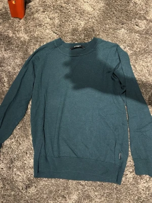 Mörkgrön tröja från Tiger of Sweden - Snygg mörkgrön tröja från Tiger of Sweden med rund hals och långa ärmar. Tröjan är tunnstickad och har diskret logotypdetalj vid nederkanten. Perfekt för lager-på-lager och stilrena outfits.
