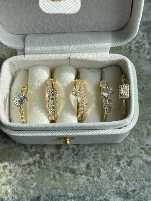 Set med fem guldpläterade och justerbara ringar - Säljer ett set med fem eleganta ringar i Copper pläterade i 18k för bättre hållbarhet mot oxidering och missfärgning ( ej stämplade).. Alla dekorerade med gnistrande klara stenar i olika former och storlekar. Ringarna har både klassiska och moderna designer, vissa med större stenar och andra med rader av mindre stenar. Perfekt för dig som gillar att mixa och matcha smycken. Alla ringar har öppning och kan justeras i storleken efter fingerstorleken.