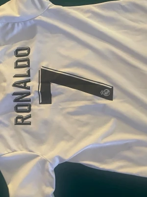 Vit Real Madrid Ronaldo tröja - Säljer en vit fotbollströja från Real Madrid med Ronaldo och nummer 7 tryckt på ryggen. Tröjan har korta ärmar och är tillverkad i ett lätt och ventilerande material, perfekt för träning eller match. Klassisk design med klubbens logga på siffran.