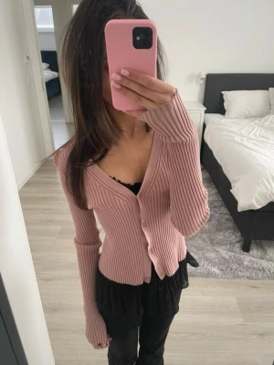 Rosa ribbad kofta  - Rosa kofta med ribbad struktur och v-ringning. Koftan har långa ärmar och knäpps framtill med matchande knappar. Passar perfekt till lager-på-lager och ger en snygg siluett. Materialet är mjukt och stretchigt.