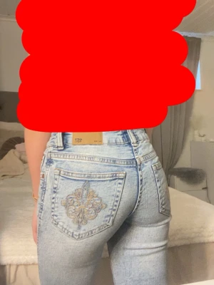 Ljusblå broderade jeans från 157 - Säljer ett par ljusblå jeans från 157 med snyggt guldbroderi på bakfickorna. Jeansen har lågmidja, bootcut och klassisk femficksmodell. Materialet är denim i bomull och de har en cool vintagekänsla. Perfekta för dig som vill sticka ut lite extra!