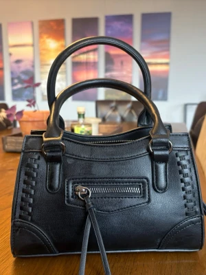 Svart handväska med dubbla handtag - Snygg svart handväska i konstläder med dubbla handtag och dekorativa sömmar. Väskan har en liten dragkedjeficka framtill och stilrena metallringar som detaljer. Perfekt storlek för det viktigaste och en clean look som passar till många outfits.