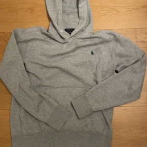 Grå hoodie från Polo Ralph Lauren - Snygg grå hoodie från Polo Ralph Lauren med klassisk huva och känguruficka. Tröjan har det ikoniska lilla blå logobroderiet på bröstet och är gjord i mjuk bomullsblandning. Perfekt för en avslappnad och stilren look.