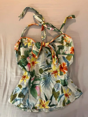 Blommig linnetopp med knyt - Säljer en somrig linnetopp med färgglatt blommönster i grönt, orange, rosa och gult. Toppen har knytband i nacken och framtill samt smockad rygg för skön passform. Perfekt för varma dagar och ger en fräsch vibe till din outfit.