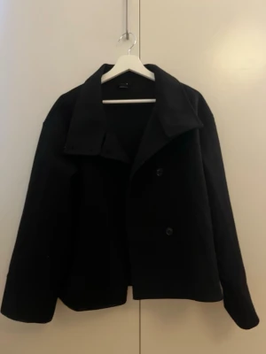 Svart kort rock från Gina Tricot - Stilren svart kort rock från Gina Tricot med bred krage och dubbelknäppning framtill. Klassisk och enkel design som passar till många olika looks. Tillverkad i ett tjockare tyg.