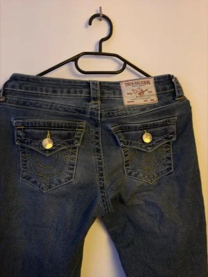 True Religion bootcut jeans blå - Säljer ett par blå bootcut jeans från True Religion med ikoniska fickor bak, dekorerade med kontrastsömmar och stora metallknappar. Klassisk femficksmodell i denim med snygg tvätt och läderpatch i midjan. Perfekta för en avslappnad och trendig look. Så gott som nya