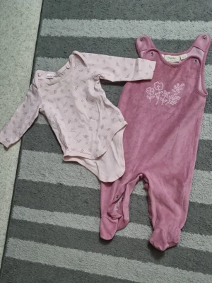 Rosa body och overall  - Säljer ett set med en ljusrosa långärmad body med små blommönster och en mörkrosa overall med broderade blommor framtill. Båda plaggen har tryckknappar för enkel påklädning och är gjorda i mjukt material, perfekt för bekväma dagar.