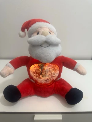 En stor jultomte med snöglob och belysning - Mysig jultomte i plysch med inbyggd snöglob i magen. Inuti globen finns en liten gatlykta som lyser, sjunger och snö som virvlar runt en miniatyrgran. Perfekt som dekoration till julen. Tomten är röd och vit med svart på fötterna och har ett glatt ansikte.