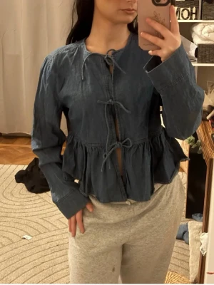Blå jeansblus med knyt framtill - Säljer en blå jeansblus med volang nedtill och tre dekorativa knytband framtill. Blusen har rund halsringning och långa ärmar. Materialet är denimliknande bomull som ger en avslappnad men trendig look. Köpt för 1000. Passar även S