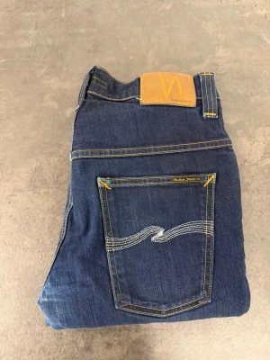 Mörkblå jeans från Nudie Jeans W32 L34 - Snygga mörkblå jeans från Nudie Jeans med klassisk femficksmodell och kontrastsömmar. Bakfickan har den ikoniska vågiga sömmen och läderpatch med logga i midjan. Jeansen är raka i modellen och tillverkade i denim av bomull.