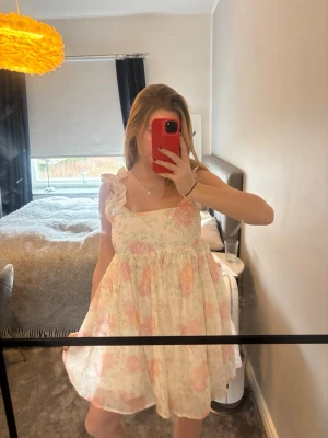 Blommig kort klänning - Söt kort klänning med blommigt mönster i ljusa toner av rosa och beige. Klänningen har volangdetaljer på axelbanden och en luftig passform. Perfekt för varma dagar och somriga tillfällen. Endast använd 1 gång och är som ny