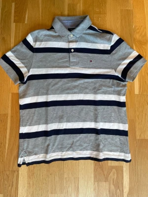 Vintage Retro Tommy Hilfiger Piké 2000s - Klassisk pikétröja från Tommy Hilfiger med breda marinblå och vita ränder på grå botten. Tröjan har krage, två knappar och korta ärmar. Liten logga broderad på bröstet. Perfekt för en avslappnad och stilren look.