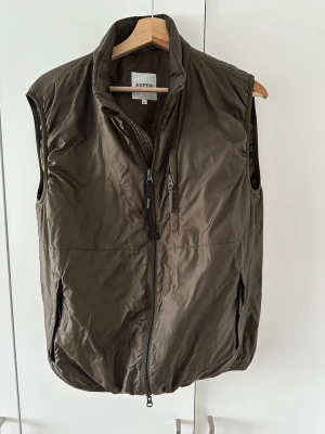 Olivgrön Aspesi Jil väst - Snygg olivgrön dunväst från Aspesi, i modellen Jil. Västen har två sidofickor med dragkedja och en bröstficka. Tillverkad i ett lätt material som håller värmen utan att kännas klumpig, perfekt för lager på lager eller över en skjorta på svalare sommardag.