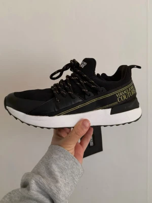 Svarta Versace Jeans Couture sneakers i storlek 37 - Snygga svarta sneakers från Versace Jeans Couture med vita sulor och gulddetaljer. Skorna har svarta och guldfärgade snören, logga på sidan och mesh- samt läderdetaljer för en sportig och trendig look.