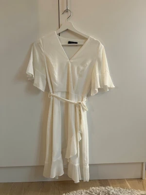 Vit omlottklänning från Sisters Point - Supersöt vit omlottklänning från Sisters Point med volangdetaljer och knytband i midjan (som kan tas bort). Klänningen har korta, fladdriga ärmar och v-ringning. Perfekt midsommar eller studentklänning! Klänningen är 100% polyester, så väldigt luftig och skön.