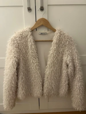Krämvit teddyjacka från Gina Tricot - Supersnygg ljus pälsjacka från Gina Tricot med lång luddig päls och öppen front. Jackan har en croppad passform och långa ärmar, perfekt för att höja din vinterstil. Färgen är krämvit och materialet känns mjukt och gosigt.