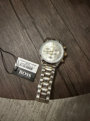 Silverfärgad klocka från Hugo Boss - Stilren klocka från Hugo Boss i silver med gulddetaljer. Klockan har en rund urtavla med tre mindre subdials och datumvisning. Länken är i metall och matchar den eleganta designen. Perfekt accessoar för dig som gillar klassisk och modern stil.