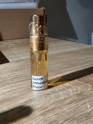 Essence Strawberry Eclair parfym 3ml, SPLITT - En söt parfym med doft av jordgubb och eclair från Essence. Flaskan är genomskinlig med guldfärgat lock och detaljer, och har en handskriven etikett. Perfekt för dig som gillar fruktiga och bakverksinspirerade dofter.