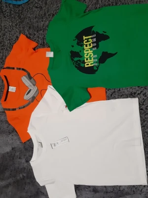 Helt nya . 50 kr/st. 3-pack t-shirts från Lindex - Säljer ett 3-pack t-shirts från Lindex i olika färger och tryck. En grön med texten 'Respect Our Home' och världskarta, en orange med hörlursmotiv och en klassisk vit t-shirt. Alla har rund hals och korta ärmar. Perfekta för en avslappnad och cool stil.