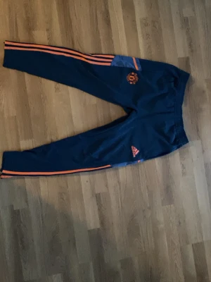 Manchester United träningsbyxor Adidas - Mörkblå träningsbyxor från Adidas med Manchester United-logga och orangea detaljer. Byxorna har smal passform, elastisk midja och klassiska Adidas-ränder längs benen. Perfekta för fotbollsträning eller gymmet.