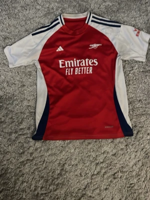 Arsenal matchtröja Adidas röd/vit - Snygg Arsenal matchtröja från Adidas i rött och vitt med mörkblå detaljer. Tröjan har klubbens klassiska kanon-logga broderad på bröstet och Emirates Fly Better-tryck framtill. Tillverkad i lätt och ventilerande material, perfekt för fotbollsträning.