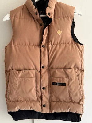 Beige dunväst från Canada Goose - Snygg beige dunväst från Canada Goose med två stora framfickor, tryckknappar och hög krage. Västen har en diskret logga med lönnlöv på bröstet och svart innerfoder. Perfekt för lager-på-lager och håller dig varm under kyliga dagar.