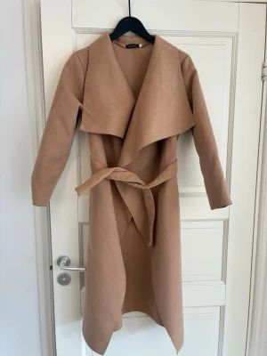 Beige lång kappa med knytskärp - Stilren beige kappa med lång passform och knytskärp i midjan. Kappan har bred krage och är tillverkad i ett mjukt tyg som ger ett snyggt fall. Perfekt för dig som vill ha en klassisk och trendig look till hösten eller våren.