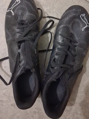 Svarta Puma fotbollsskor med snörning - Säljer ett par svarta fotbollsskor från Puma med vita loggor och diskreta prickmönster. Skorna har klassisk snörning och är tillverkade i syntetmaterial som ger bra grepp och komfort på planen. Perfekta för dig som vill ha stilrena och funktionella skor.