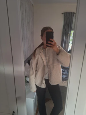 Beige teddyjacka med knappar - Mysig beige teddyjacka med knäppning framtill och två stora fickor. Jackan har en rak passform och är tillverkad i ett fluffigt teddy-material som ger en avslappnad och trendig look. Perfekt för kyliga dagar och enkel att matcha med olika outfits.