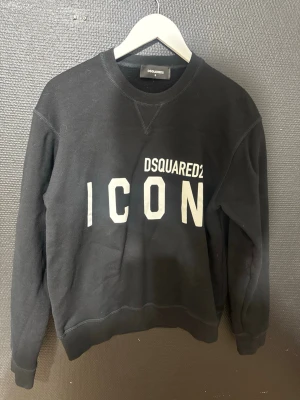 Svart sweatshirt Dsquared2 ICON - Cool svart sweatshirt från Dsquared2 med stor vit 'ICON'-logga framtill. Tröjan har rund halsringning, ribbade muddar vid ärmslut och nederkant samt en avslappnad passform. Perfekt för en trendig streetwear-look.