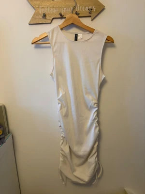 Vit omlottklänning från H&M - Säljer en vit ärmlös klänning från H&M med snörning på sidorna som ger en snygg omlott-effekt. Klänningen är kort och har en enkel, modern design som passar perfekt för sommarens alla tillfällen.